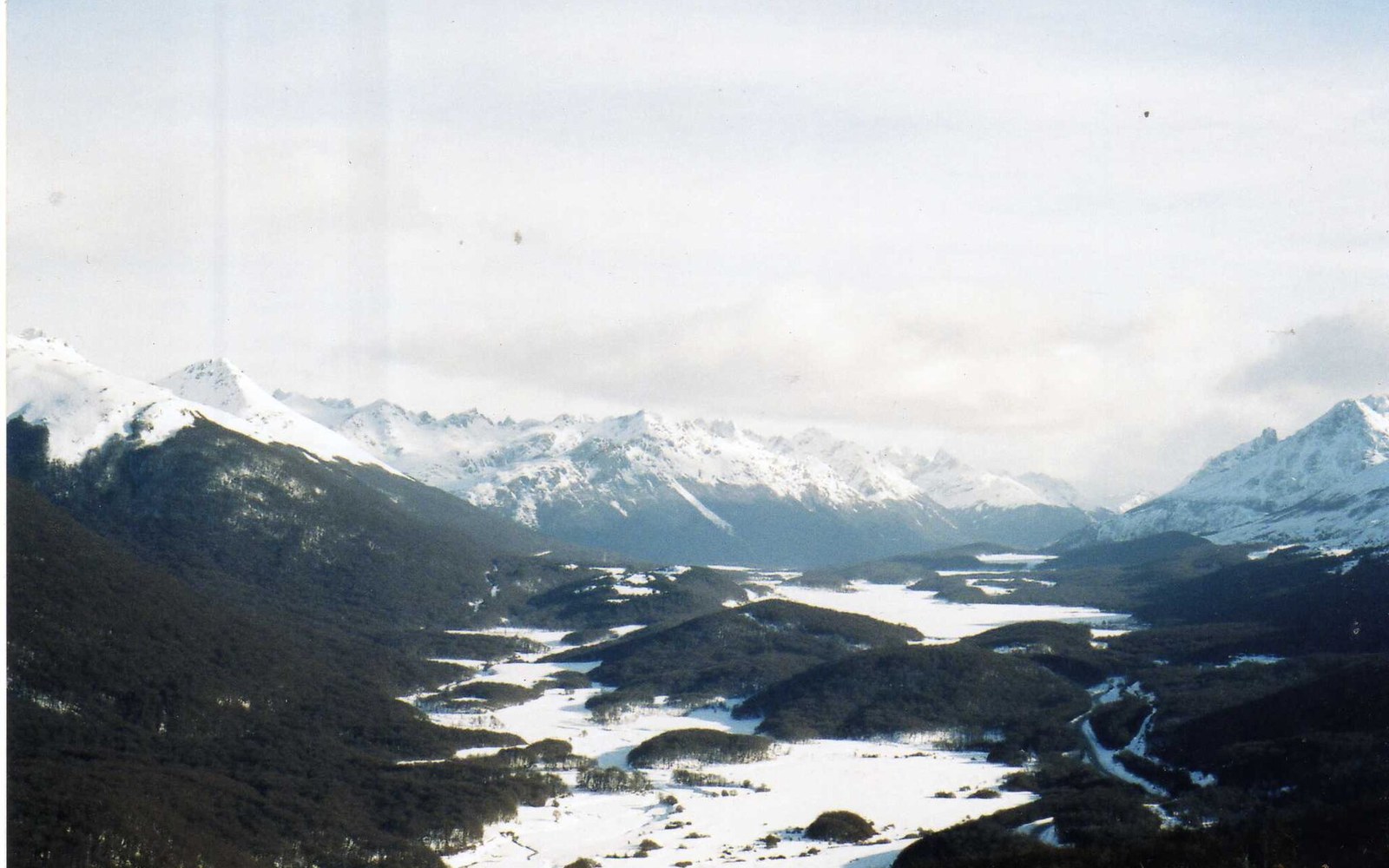 A low-treeline ski slope under flat grey southern light, lift towers running up a bare ridge, the sea visible in the distance